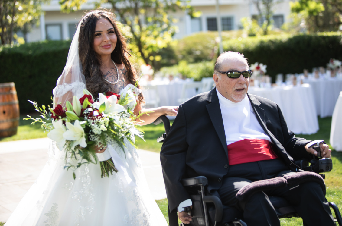 A bride in a while dress is escorted down the aisle by a man in a wheelchair